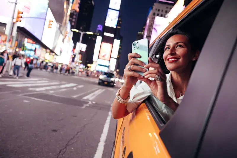 Mujer asomada por la ventanilla de un taxi tomando una foto, con Times Square iluminado al fondo.