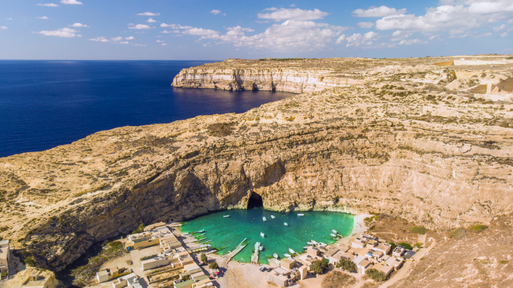 A photograph of a natural pool in the inland sea in Id Dwejra on Gozo, an island forming part of Malta. In the background, yellowy brown coloured sea cliffs stretch into the distance against a blue sky with white clouds. To illustrate a blog post entitled 'where is hot in October in Europe?'