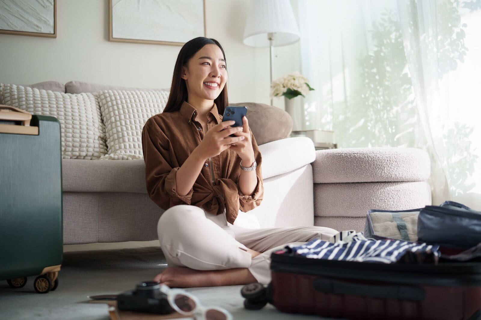 Woman holding up her phone and smiling, with a packed suitcase on the floor in front of her