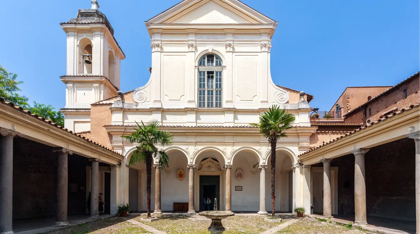View of the courtyard and fountain in front of the white facade of the Basilica of San Clemente in Rome.