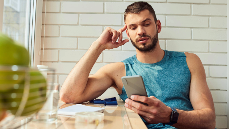 A photograph with a man in the foreground looking at his phone and looking confused or like he is trying to figure something out. He is wearing a sleeveless t-shirt in a relaxed setting, perhaps a holiday rental apartment or a hostel. This is an image to illustrate a blog post entitled 'Unlimited eSIM: Plans, Benefits, and How It Works'. 