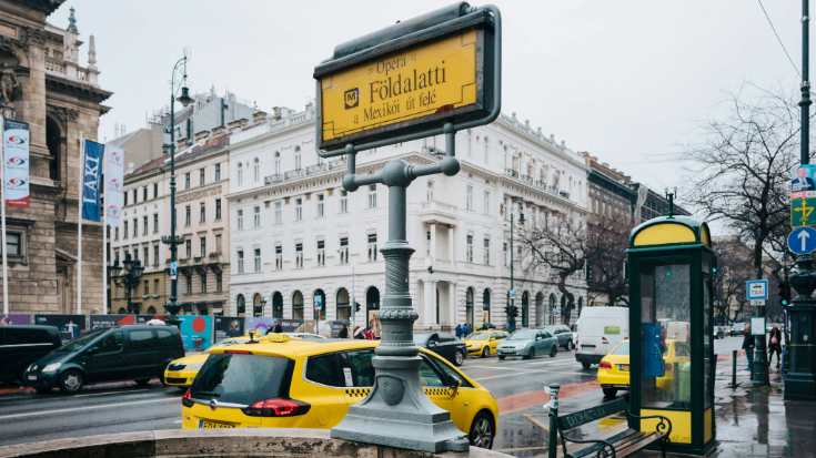 A photograph os the Foldalatti metro stop sign in Budapest in the foreground. In the background is a city scene, showing the city's grand sand-coloured buildings, some parked taxi cabs which are coloured yellow with black squared running along the side of them. To illustrate a blog post entitled 'The Ultimate Sziget Festival Guide.'
