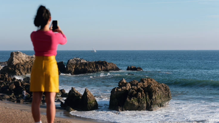 A photograph with a woman in the foreground with her back to the camera. She is slightly out of focus, but she is holding a mobile phone up to take an image of the scene in the background. She has black hair in a ponytail, is wearing a bright yellow knee-length skirt and a pink t-shirt and is standing on a beach. The scene in front of her is of a beautiful ocean shore, with clear blue skies, waters and textured rocks. This is an image to illustrate a blog post entitled 'Unlimited eSIM: Plans, Benefits, and How It Works'. 