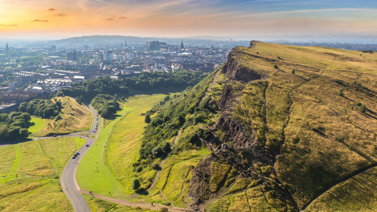 A view over Edinburgh including the green hill of Arthur's Seat, showing the cityscape as the sun sets.