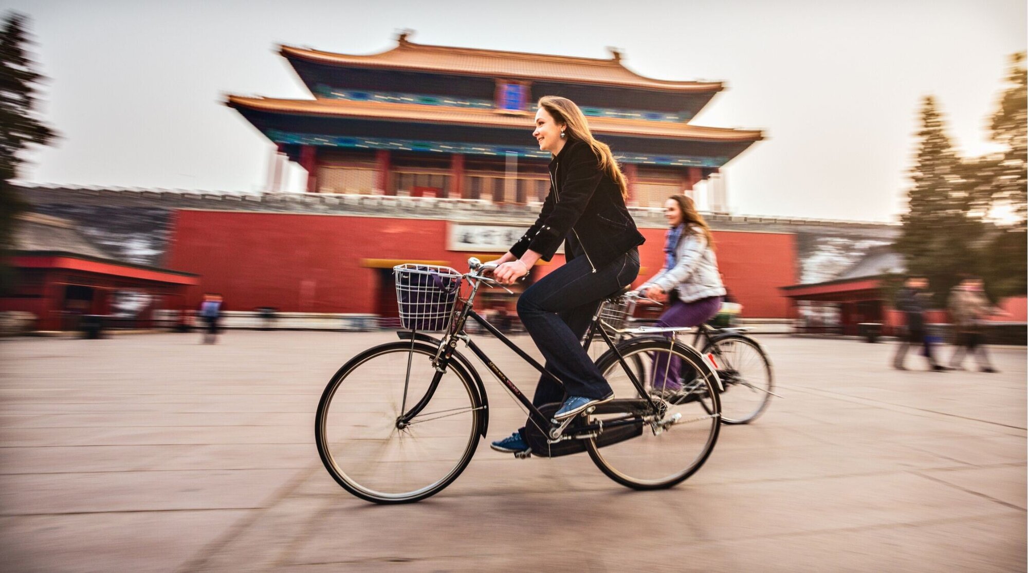 Chip internacional china: Duas mulheres andam de bicicleta em frente a um prédio tradicional chinês de arquitetura histórica, em uma cena urbana cultural.