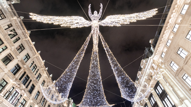 A photograph taken from the ground looking upwards to the sky at night time in the dark, looking down is a light display in the shape of an angel, made using white LED bulbs. These are Christmas lights on display in London's Regent street at Christmas time. To illustrate a blog post entitled 'Things to do in London in December'. 