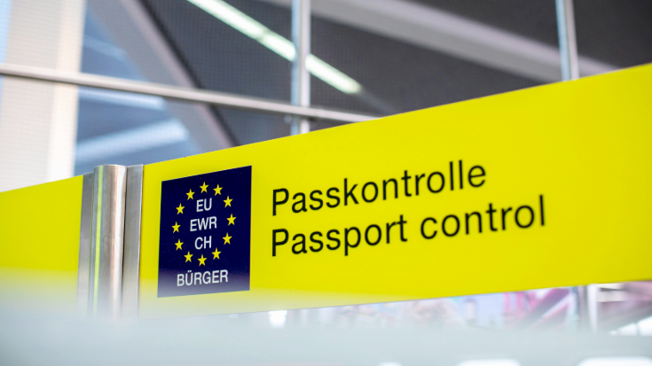 A photograph of a bright yellow sign in an airport, which states 'Passport Control' and 'Passkontrolle' across it in black letters. There is also a blue graphic next to it featuring yellow stars and stating 'EU/EWR/CH/BURGER'. To illustrate a blog post entitled 'What's the Difference Between the EES and ETIAS? A Guide for Brits.'