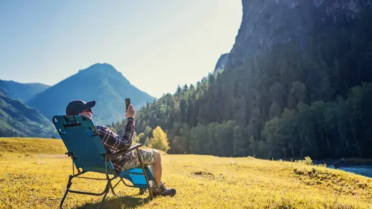 Person sitting in a camping chair in a mountain meadow, using a smartphone to activate a travel eSIM. 
