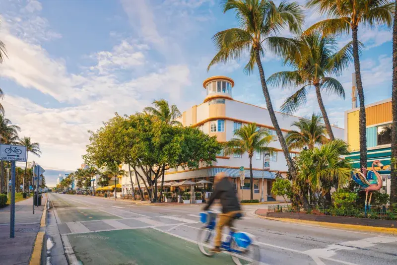 Ocean Drive en Miami Beach por la mañana, con palmeras, edificios art déco y una bici pasando.