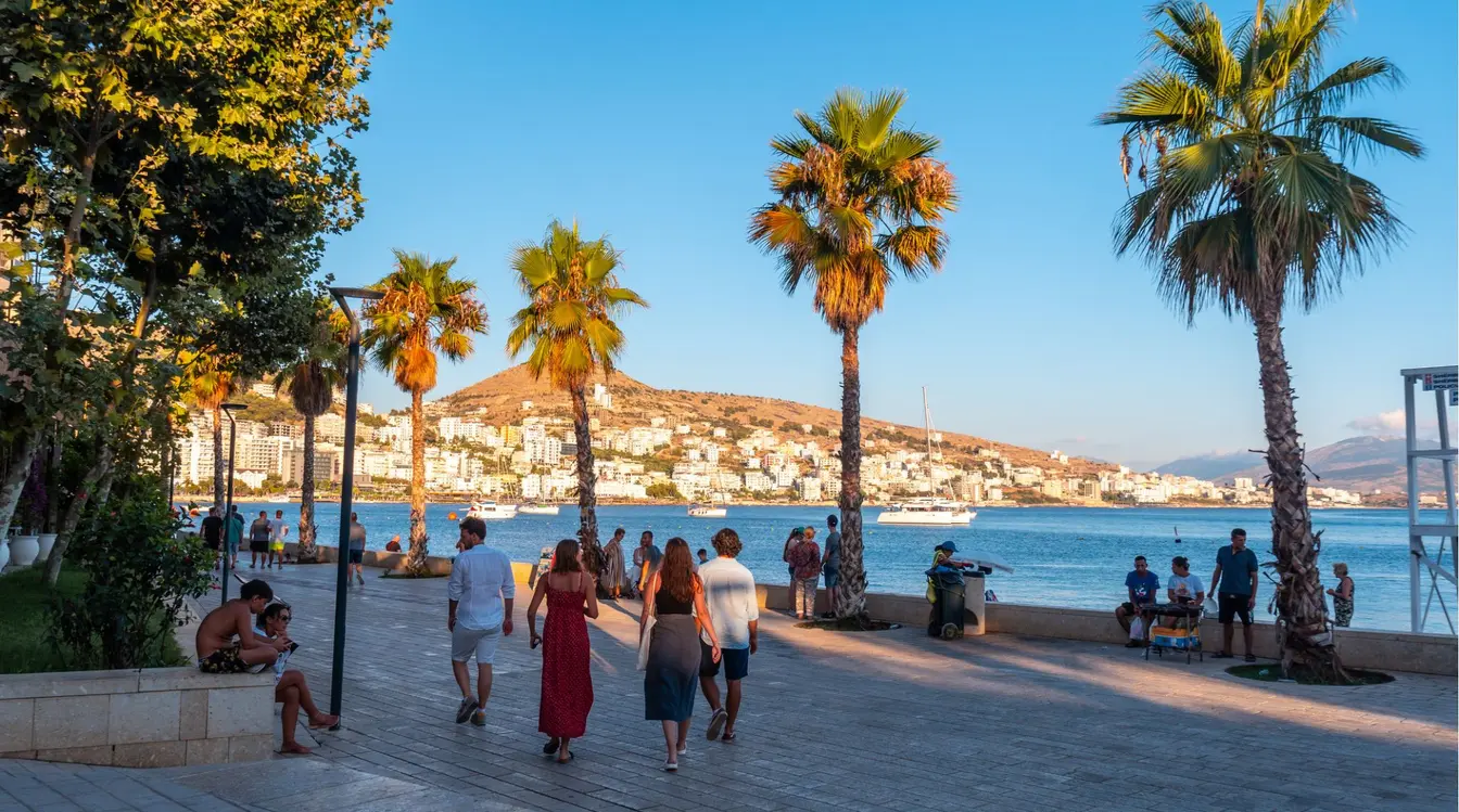Turistas caminhando na orla de Saranda, cidade costeira da Riviera Albanesa, com palmeiras e o mar Mediterrâneo ao entardecer.