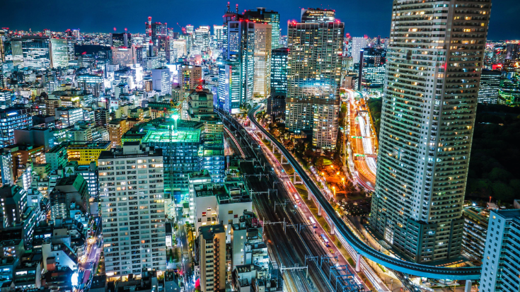 A night time arial photograph of Tokyo, showing busy streets below with cars going to and from and busy buildings with lights shining from office windows.