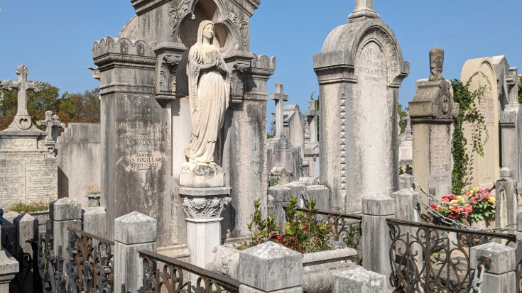 A colour photograph of Cimetière De Loyasse in Lyon, France. In the background, the sky is clear pale blue and it's a sunny day. In the foreground are tightly-packed tombstones in grey stone, decoarted with carved letters and words. Some are surrounded by spiked fences with elaborate ironwork. One of the graves is decorated with a statue of the Virgin Mary and some are weathered by dark spots, creating a beautiful, aged and dramatic effect. To illustrate a blog post entitled '10 of The World's Most Beautiful Cemeteries'.