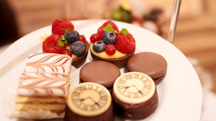 A close-up image of a selection of small bite-sized square cakes on a plate, afternoon tea in Vienna. There are blueberries and raspberries perched on top of chocolate tortes and also Vienna slices, to illustrate a blog post entitled Extreme Day Trips from London to Europe.