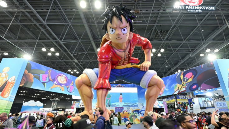 A colour photograph of the conference floor of New York Comic Con, picturing a giant-sized figure from a manga series TV show or computer game. The figure is a young man with spiked black hair. He is crouching down, staring ahead and is wearing blue denim shorts rolled at the knee, along with a read shirt.