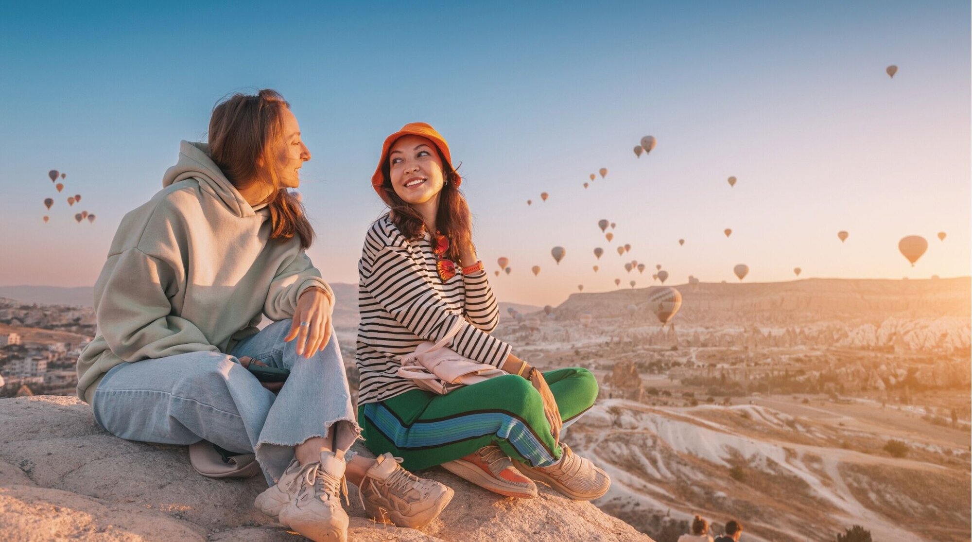 Dos amigas sentadas en un mirador miran el paisaje al atardecer, con globos aerostáticos en el cielo.