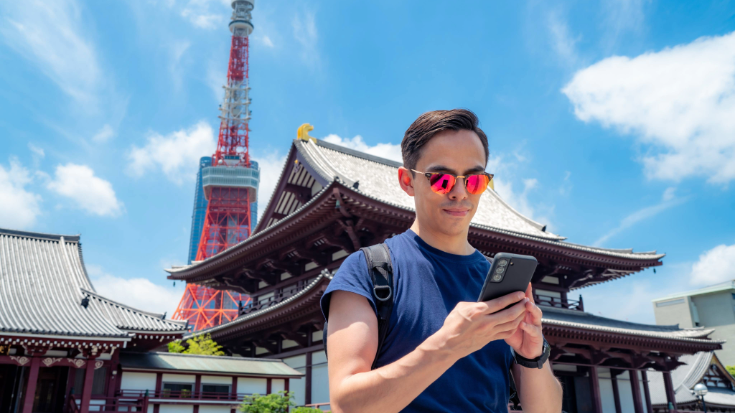 Traveller using a smartphone outside Zojoji Temple with Tokyo Tower rising behind under a bright blue sky — showing how Japan eSIM plans keep Tokyo Game Show visitors connected while exploring the city.