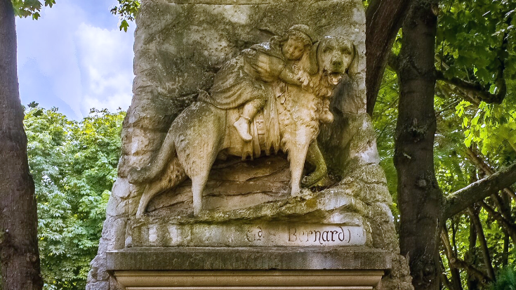 Detailed yellow sandy coloured stone relief of Barry the St. Bernard, one of history’s most famous rescue dogs, carrying a young child on his back through the snow, part of his memorial statue at the Cimetière des Chiens near Paris.