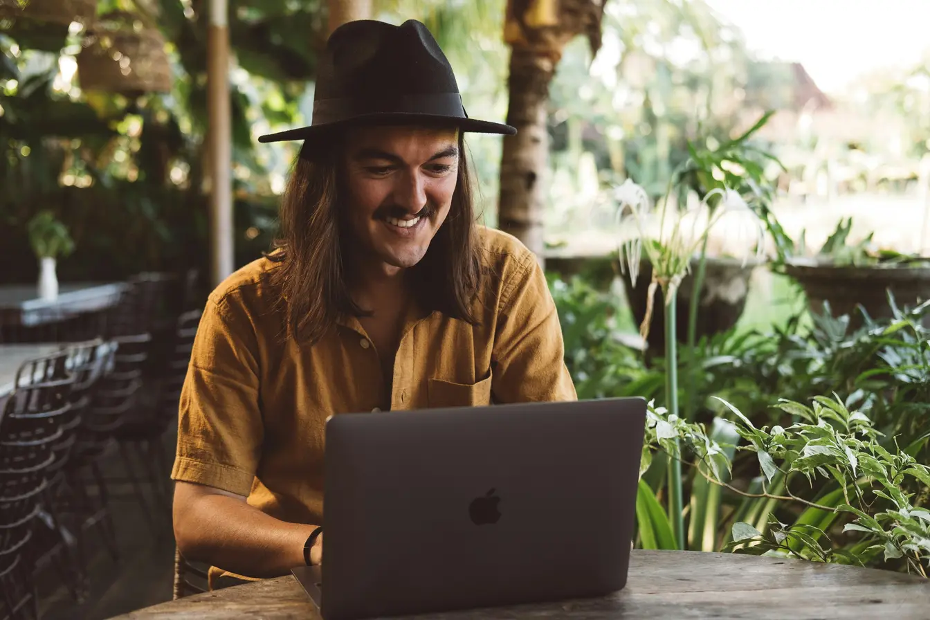 Imagen de una persona sonriente trabajando en su portátil rodeado de naturaleza para ilustrar a los nómadas digitales.