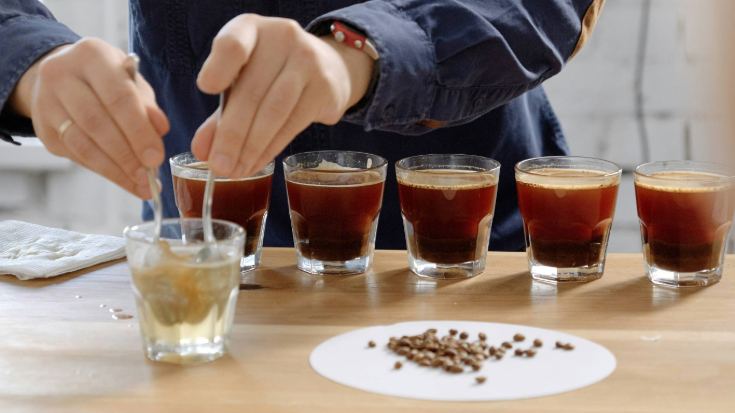 A photograph showing a close-up of a male coffee baristas hands as they mix a coffee in a small clear glass with two spoons in the foreground. In the background, five glasses filled with black coffee sit in a row. The image is an example of how a coffee cupping, or tasting session, might look, for a blog post entitled 'Which Country Has the Best Coffee Culture?.