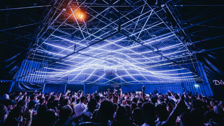 A photograph of an artistic light installation on the roof of a music venue at night. The installation is a series of slim tubes intertwining with one another to create an interesting line-based optic pattern. The lines are a cool blue light while the background is dark. There are crowds of people below the ceiling, dancing with their hands held in the air. To illustrate a blog post entitled 'The Ultimate Sziget Festival Guide.' 