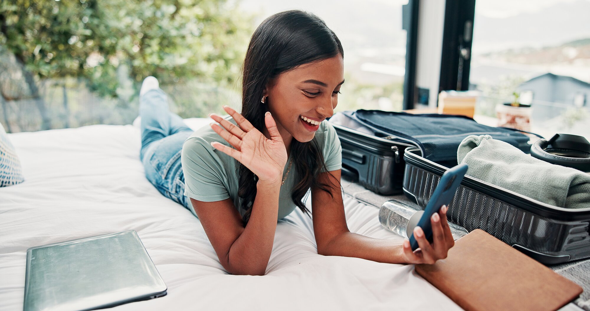 Donna felice che saluta durante una videochiamata dal telefono in una stanza d’hotel, usando un’app per fare videochiamate durante un viaggio.