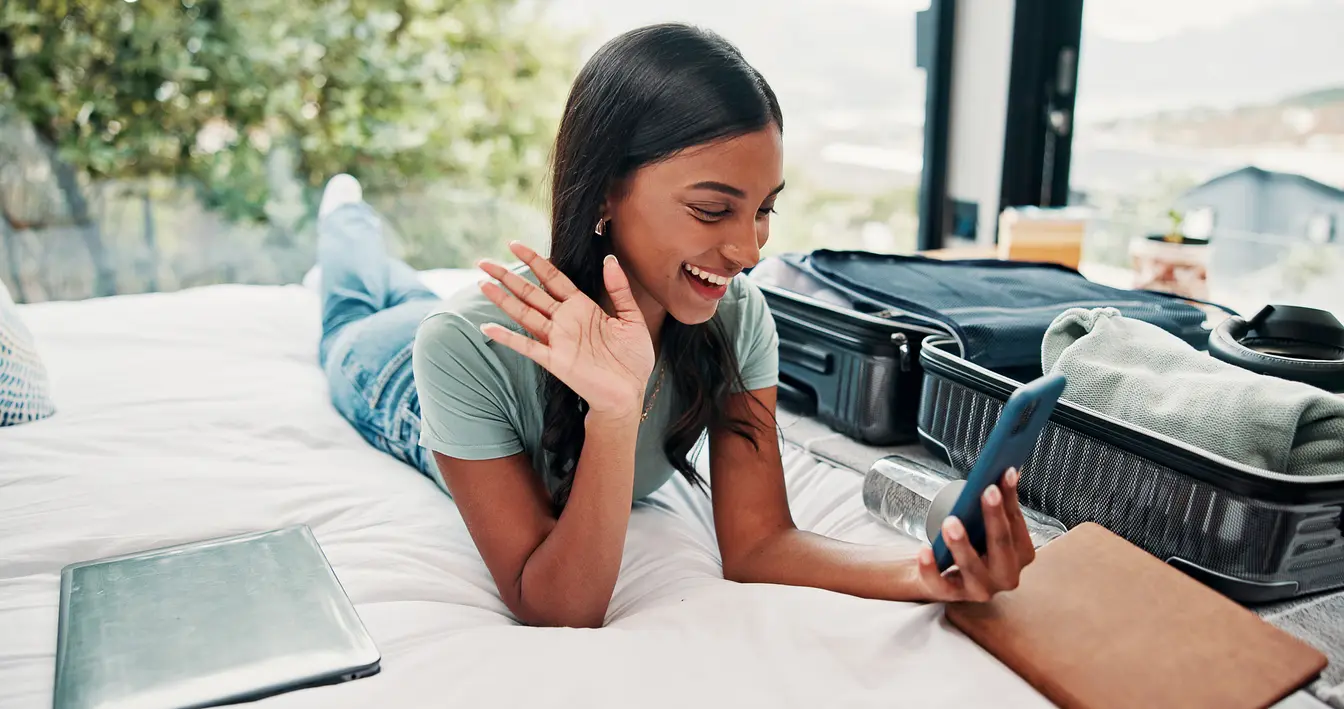 Donna felice che saluta durante una videochiamata dal telefono in una stanza d’hotel, usando un’app per fare videochiamate durante un viaggio.