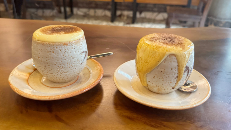 A photograph of two textured stoneware coffee mugs on matching saucers with spoons perched on them. In the background is a blurred-out cafe. The cups are overflowing with a thick yellow froth, which is egg coffee, a famous drink in Vietnam and Hanoi. for a blog post entitled 'Which Country Has the Best Coffee Culture?'