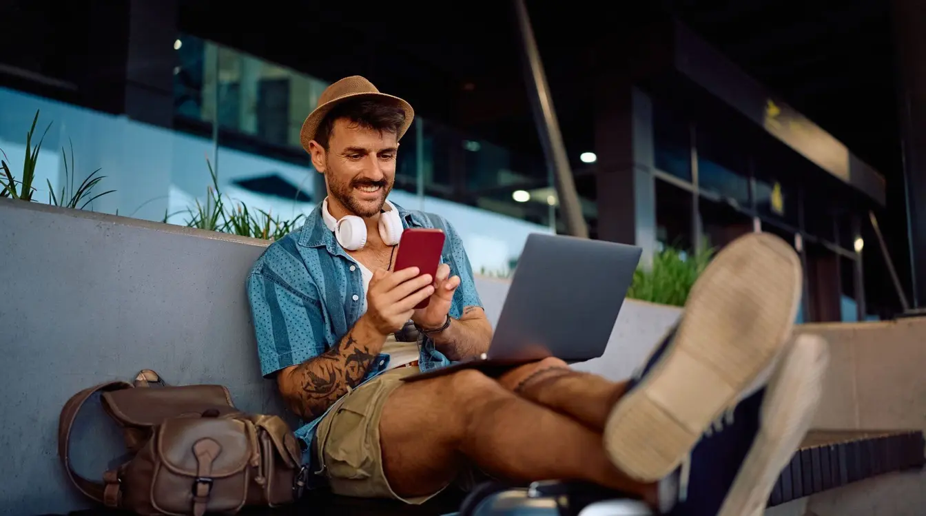 Un viajero revisa su teléfono mientras trabaja con su laptop al aire libre.
