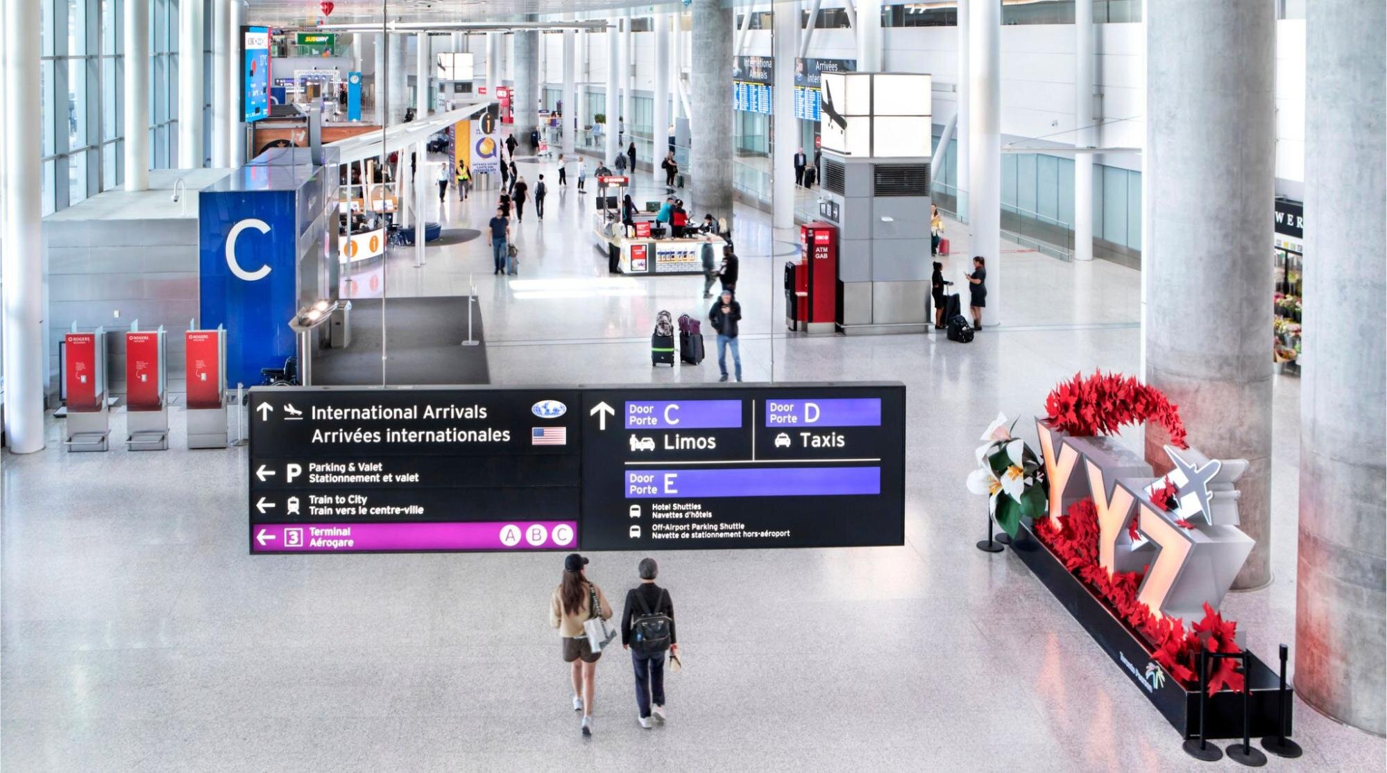  Viajantes caminhando pelo Terminal do aeroporto Internacional Toronto Pearson, em direção ao portão de Desembarque Internacional.]