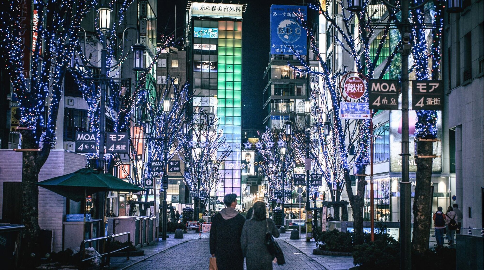 Tóquio. A imagem mostra uma rua movimentada em Kabukicho, Shinjuku, Tóquio, iluminada por numerosos letreiros de neon e luzes natalinas. A rua está cheia de pessoas caminhando, capturando a vibrante vida noturna e atmosfera urbana deste famoso distrito de entretenimento.