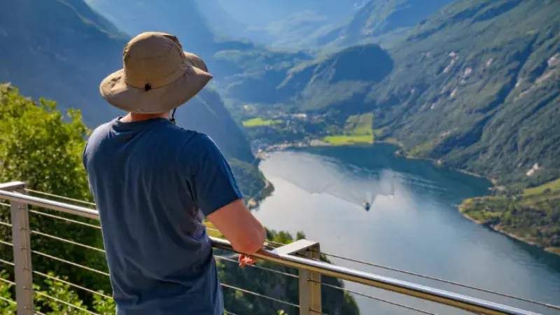 A man in a blue t-shirt, and a beige sun hat with his back to the camera, looking over a Norwegian fjord.