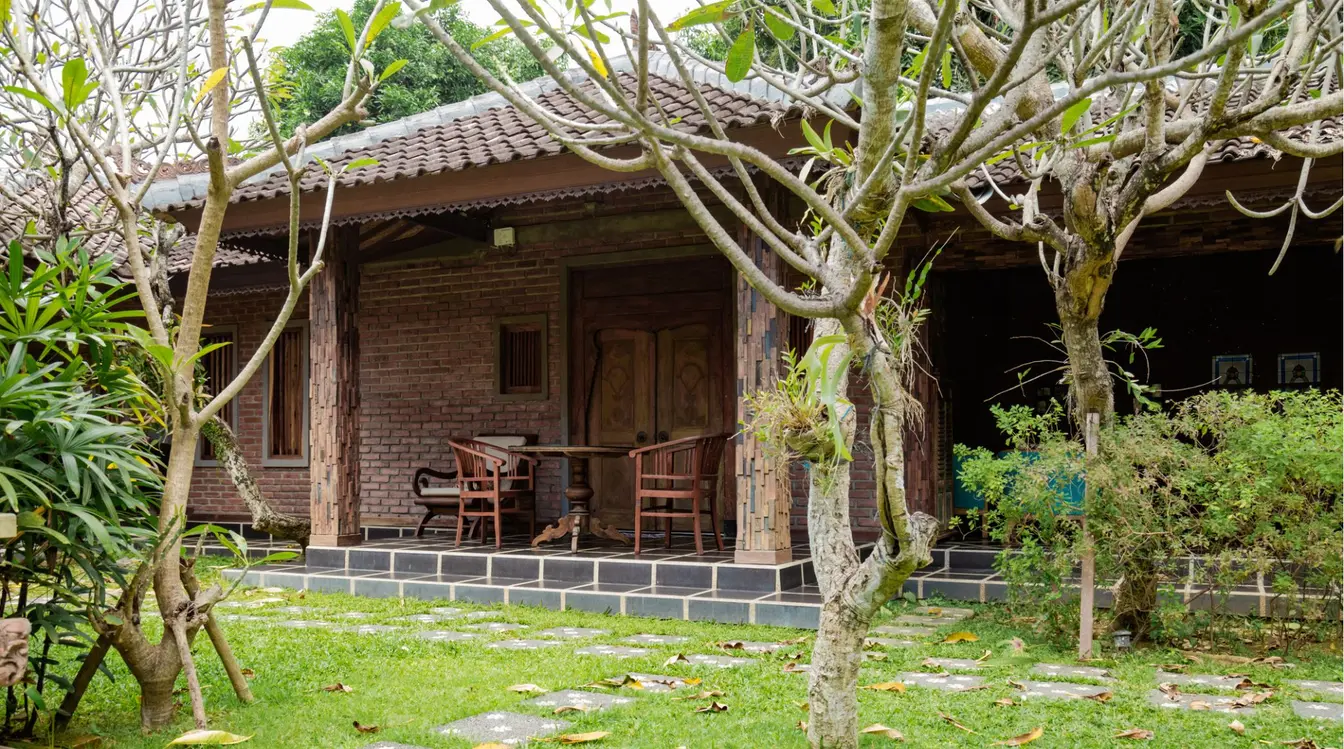 View of a courtyard and the exterior of a traditional Balinese guesthouse with a table and chairs on a covered porch.