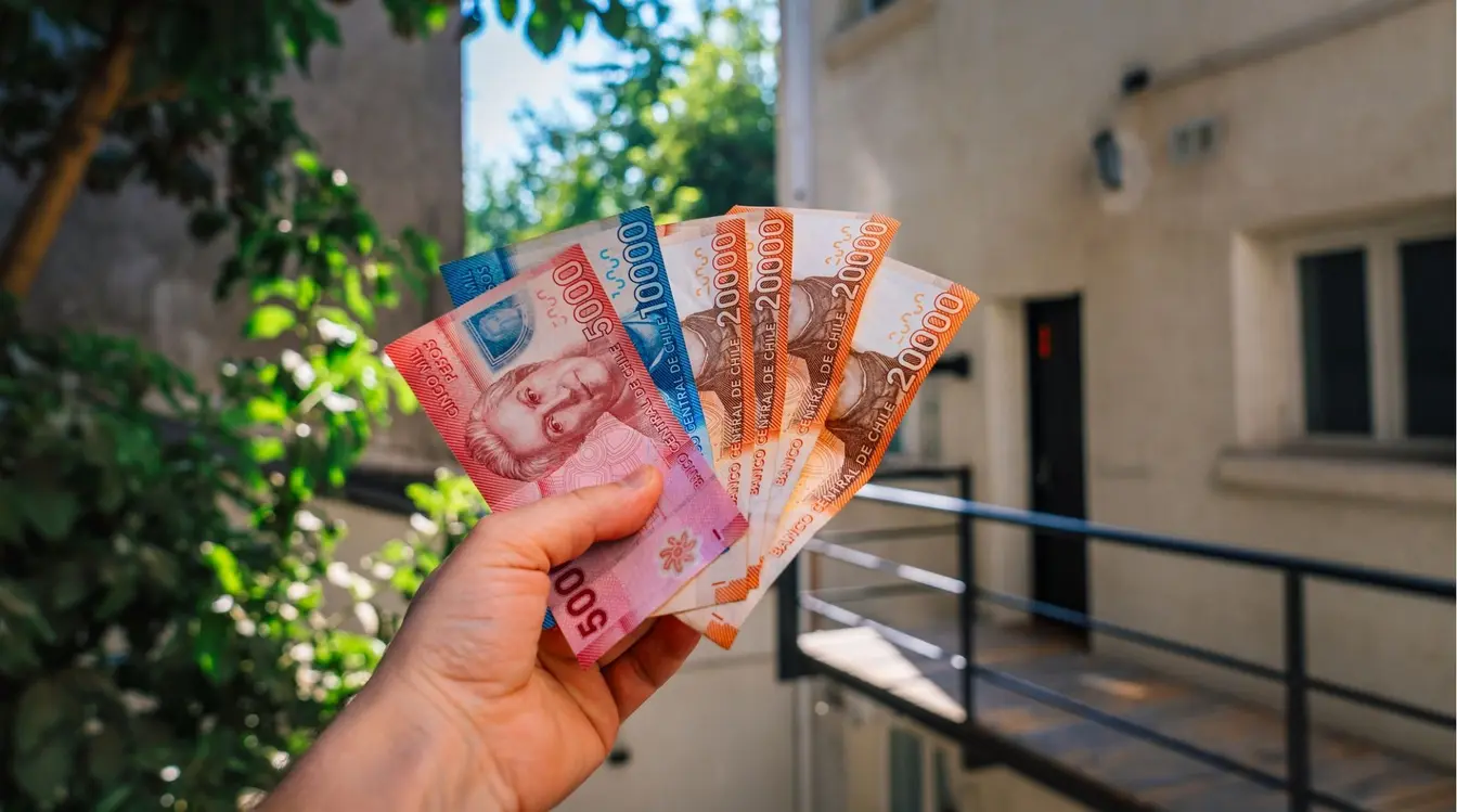 Uma mão segurando um leque de notas coloridas de pesos chilenos de diferentes valores (como 1.000, 5.000, 10.000 e 20.000), com um fundo urbano desfocado mostrando uma varanda e árvores.