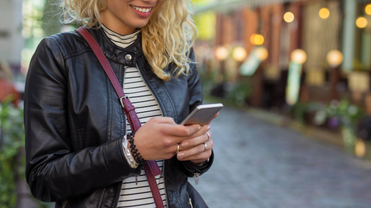 An image of a woman with curly blonde hair, a black and white horizonal striped t-shirt and a leather jacket on, holding an iPhone and smiling, while in the background is a street full of cafes and busy shops, to illustrate a blog post entitled 'Is my iPhone eSIM-compatible?'