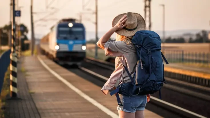 A female traveller with a blue backpack stands on a sunlit platform watching a blue train arrive, illustrating the choice of low-carbon rail travel over flying. 