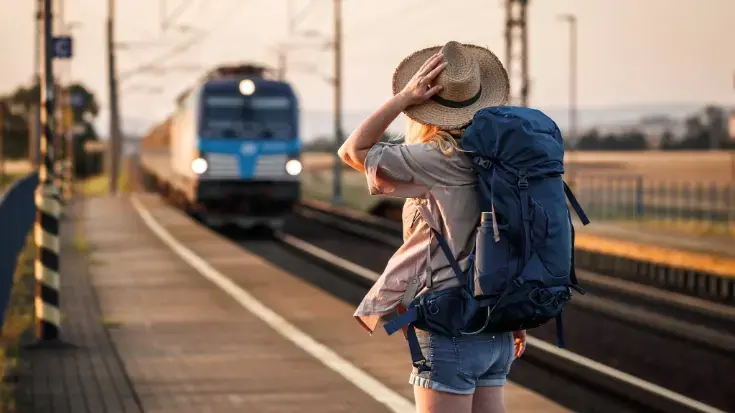 A female traveller with a blue backpack stands on a sunlit platform watching a blue train arrive, illustrating the choice of low-carbon rail travel over flying. 