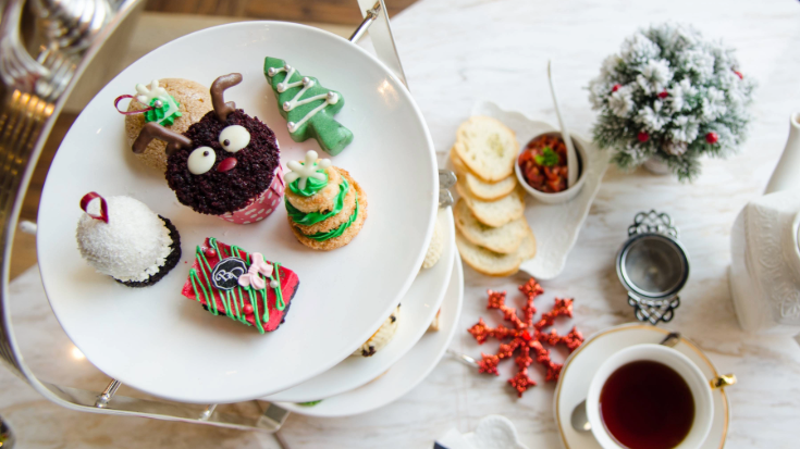 An image of some small festive afternoon tea cakes on a tiered plate at a fancy hotel, including a brown reindeer with chocolate sprinkles, a green tree with white baubles and white fondant made to look like a snow ball, to illustrate a blog post entitled 'Things to do in London in December'.