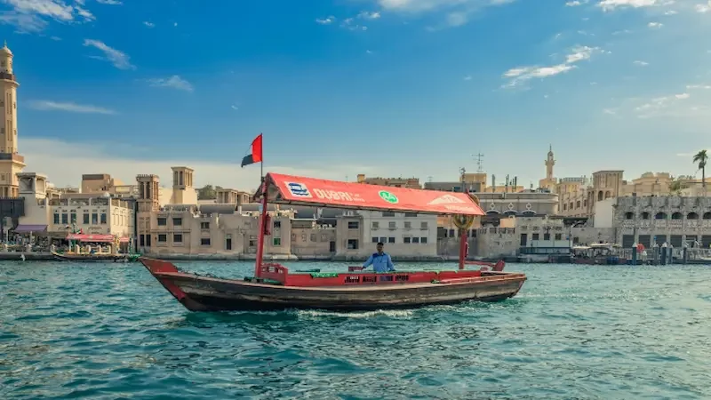 A traditional Dubai wooden ferry boat with a red canopy sailing across the blue waters of Dubai Creek. 