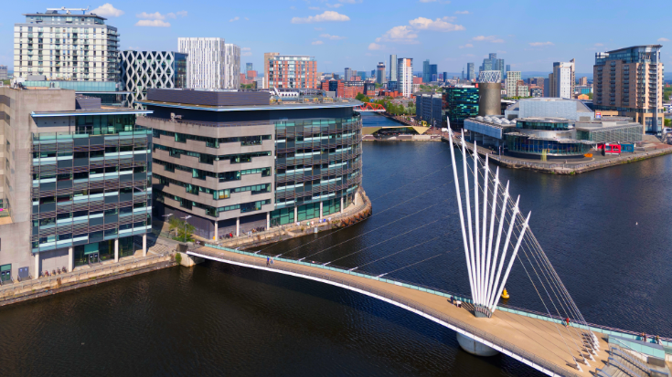 An aerial shot of Salford Quays, showing a bridge over Manchester Ship Canal and glass-fronted office and retail buildings in the frame.