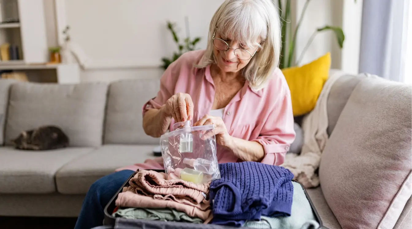 Mulher idosa de óculos e camisa rosa organiza sua mala de viagem sobre o sofá, segurando um saco plástico transparente fechado contendo pequenos frascos de líquidos.