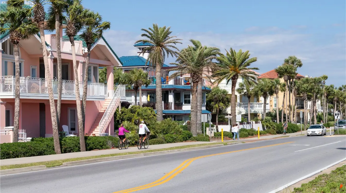 Rua residencial tranquila em Miramar, na Grande Miami, com casas de fachada colorida, palmeiras e moradores andando de bicicleta em um dia ensolarado.