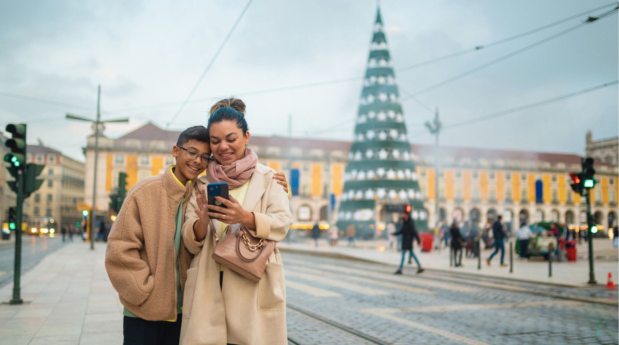 Mãe e filho usando celular com a Árvore de Natal da Praça do Comércio de Lisboa ao fundo.