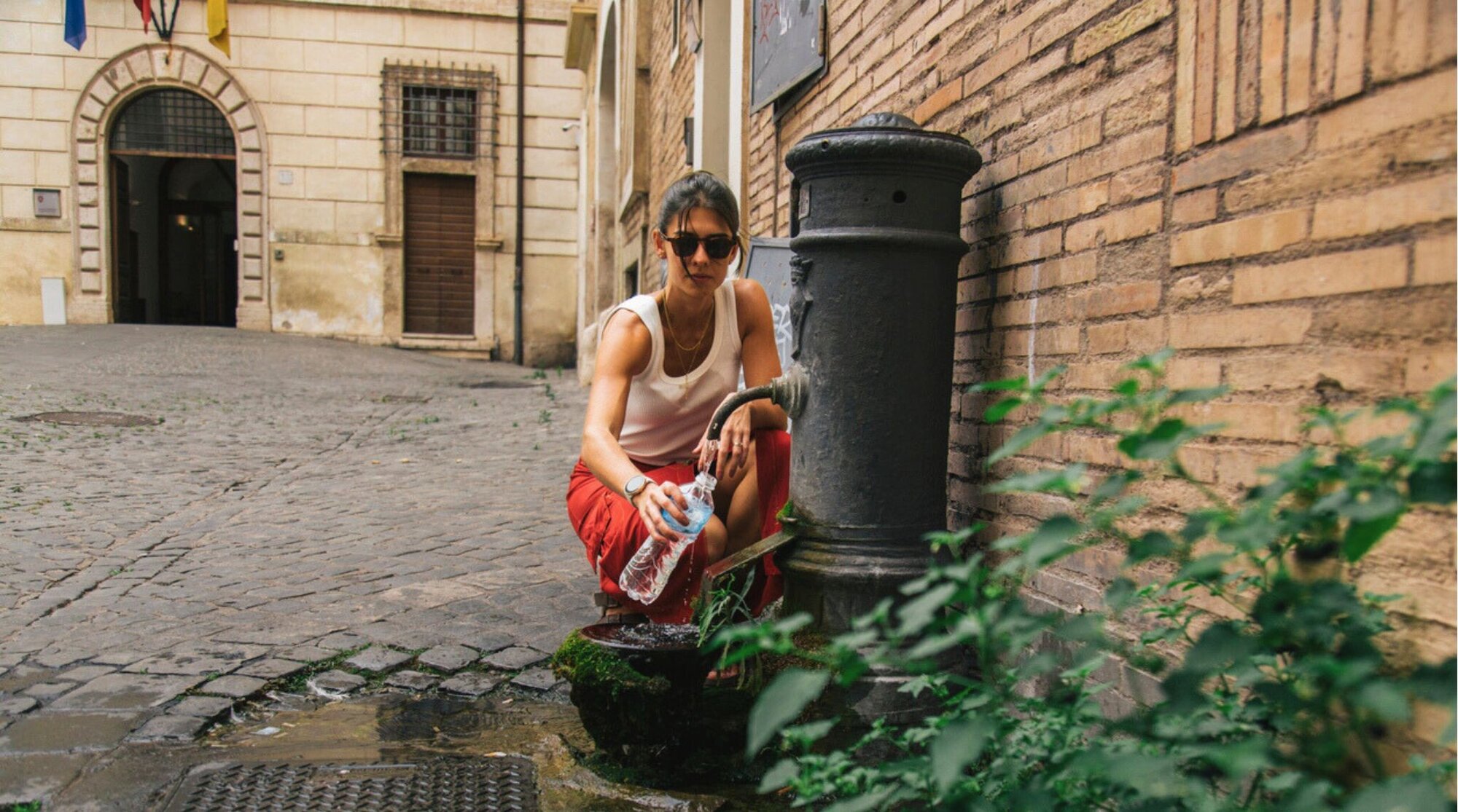 Uma mulher usando óculos de sol e roupas casuais coleta água limpa em uma garrafa plástica de uma fonte tradicional da cidade em um pitoresco beco de paralelepípedos cercado por arquitetura histórica europeia. Roma, Itália.
