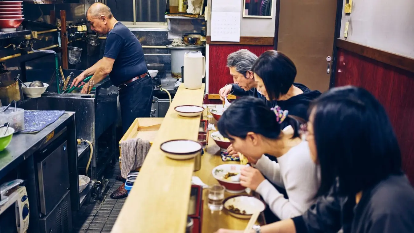 A row of people eating at a wooden counter in a traditional ramen shop in Tokyo, Japan.