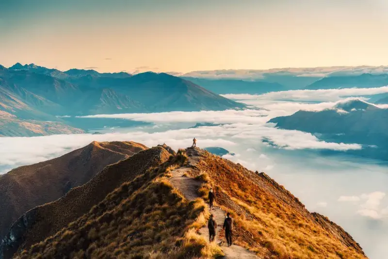 Senderistas caminando por una cresta montañosa con nubes bajas y luz suave al amanecer.