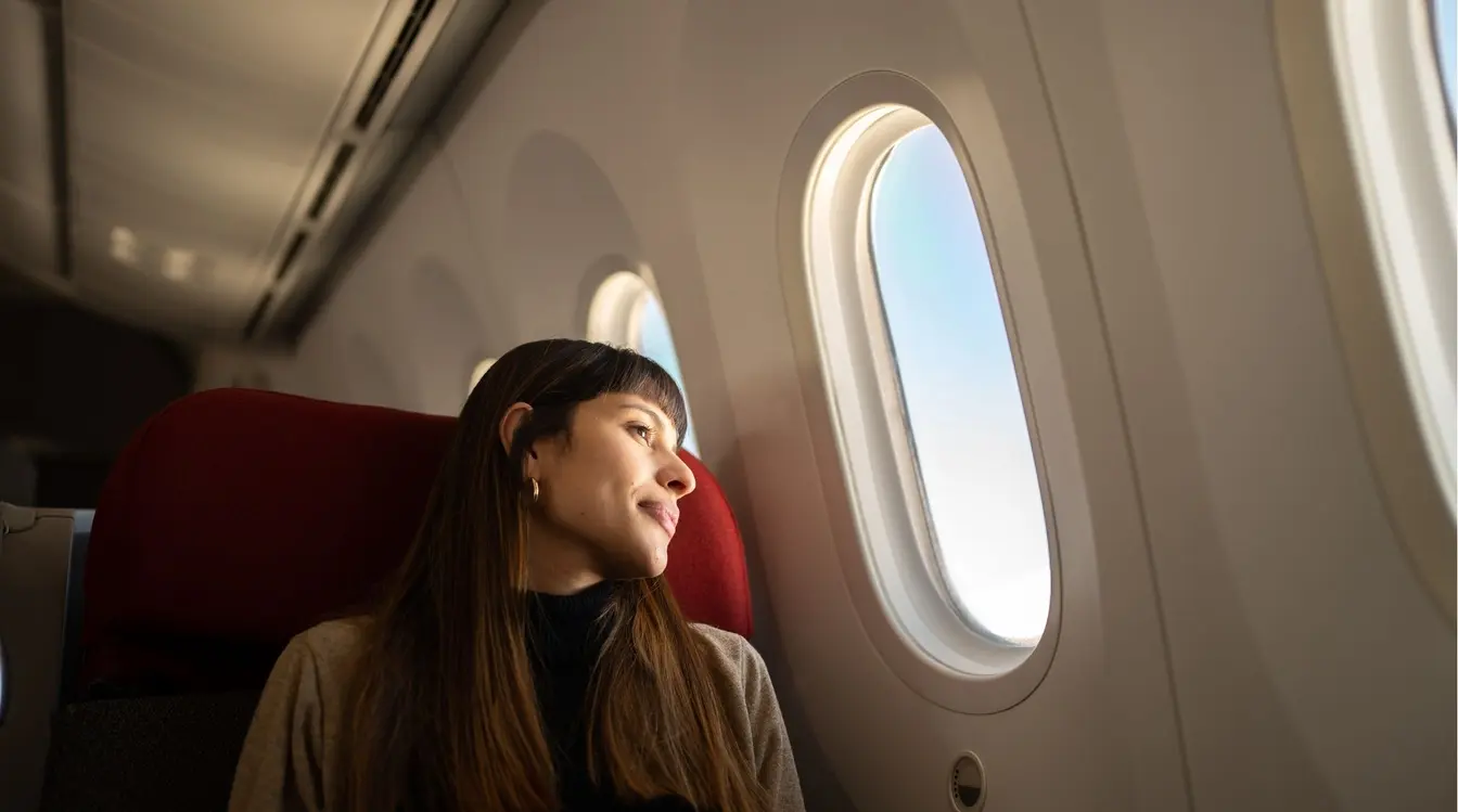 Mulher com cabelos escuros curtos sentada na poltrona de um avião, olhando tranquilamente pela janela redonda para um céu azul claro.