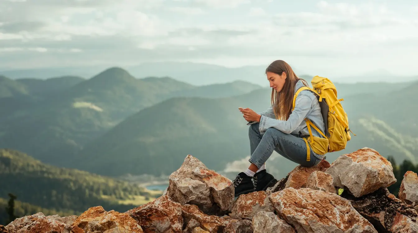 Eine junge Frau sitzt mit Rucksack in den Bergen und nutzt mobile Daten im Ausland auf ihrem Handy.