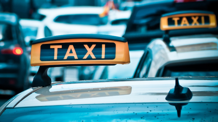 A close-up image of several taxi signs perched on the top of taxis, the signs are orange and the car are black. 