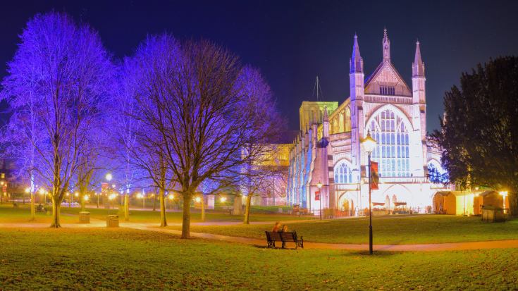 A night image of Winchester Cathedral, with a couple sitting on a bench in front of the building and surrounding trees bathed in a purple light, to illustrate a blog post entitled The Best Christmas Markets in the UK for 2024.
