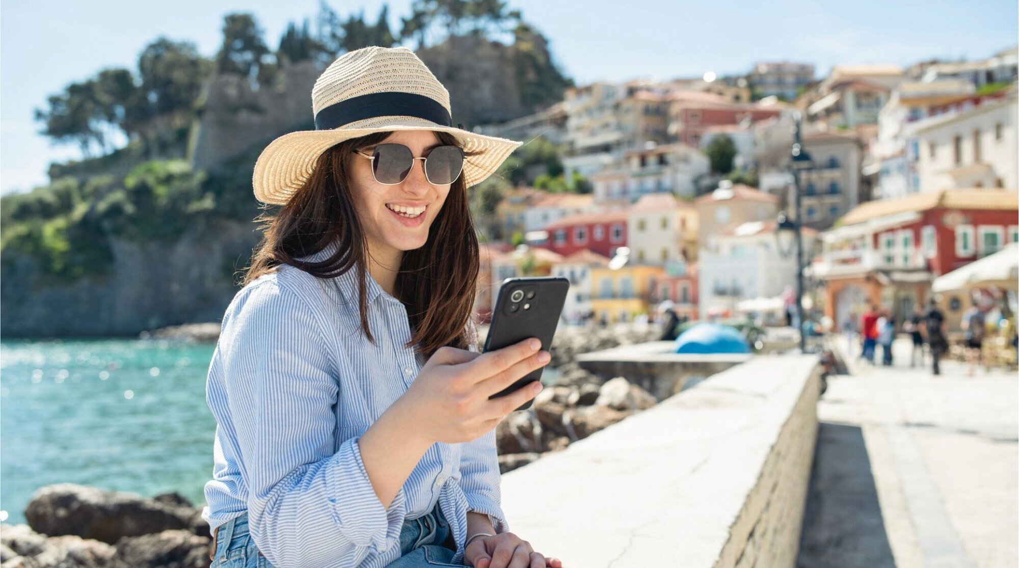 Mulher usando celular durante férias na Grécia, sentada em um café com vista para o mar.